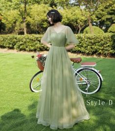 a woman in a green dress standing next to a bicycle on the grass with trees and bushes behind her