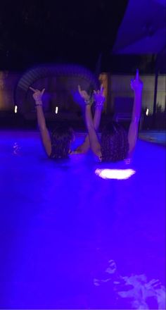 two people laying on their stomachs in the pool at night, one holding up her hand