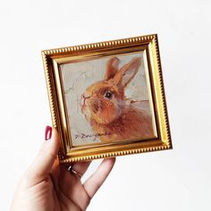 a person holding up a painting of a rabbit in a gold frame on a white wall