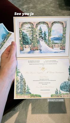 a person holding up a wedding card in front of a table with flowers and trees on it