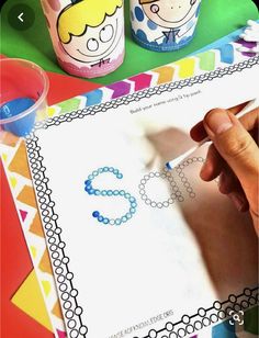 a child's hand writing the letter s on paper next to other children's cups