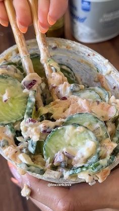 two hands holding a bowl with cucumber and chicken salad in it while another hand holds chopsticks above the bowl