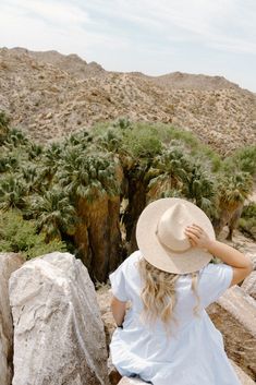 a woman in a white dress and hat sitting on rocks with palm trees behind her