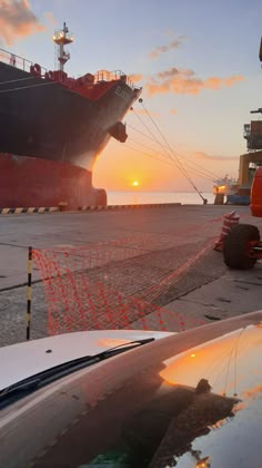 the sun is setting in front of a large cargo ship on the water with cars parked next to it