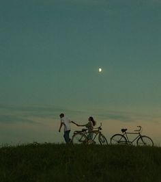 two people holding hands while standing next to their bikes on a hill with the moon in the background