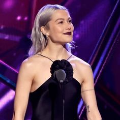 a woman standing in front of a microphone on top of a purple stage and wearing a black dress