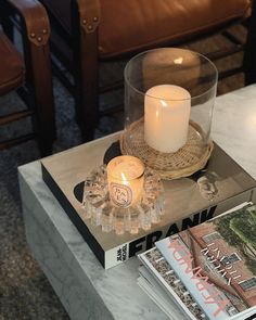 a candle and some magazines on a coffee table with chairs in the backround