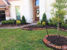 a small tree is in the middle of a flower bed by a brick wall and white house