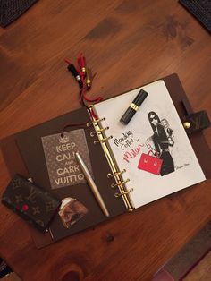 an open notebook on a wooden table with various items around it, including a pen and lipstick