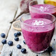 two glasses filled with blueberry smoothie on top of a wooden table next to fruit