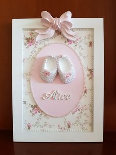 a pink and white frame with baby shoes on it's feet, in front of a brown wall