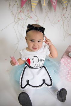 a baby sitting on the floor wearing a dress and holding a cell phone to her ear