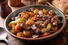 a bowl filled with meat and vegetables on top of a wooden table next to bread