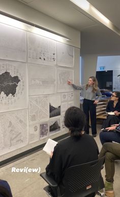 a woman standing in front of a whiteboard with maps on it and people sitting around