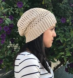 a woman wearing a knitted hat standing in front of some bushes and purple flowers