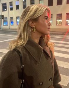 a woman standing on the street in front of a building at night wearing large gold earrings