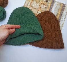 a hand holding a green knitted hat next to two balls of yarn