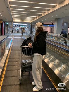 a woman standing in an airport holding a shopping cart and looking at the escalator