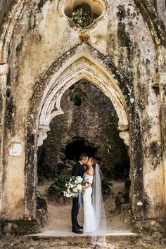 a bride and groom standing in front of an old stone archway with flowers on it