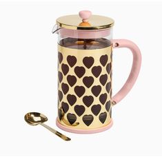 a pink and gold coffee pot with hearts on it next to two spoons,