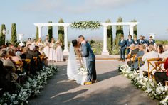 a bride and groom kissing in front of an outdoor wedding ceremony with white flowers on the aisle