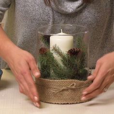 a woman is holding a candle in a glass container with pine cones and twine