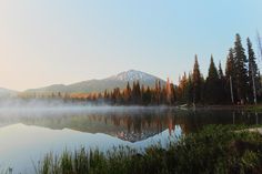 a lake surrounded by trees and fog in the air with snow capped mountains in the distance