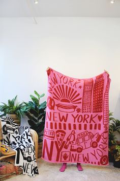 a woman standing in front of a pink blanket that has new york on it and other pillows