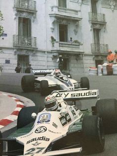 two racing cars driving down the street in front of a tall building with people watching