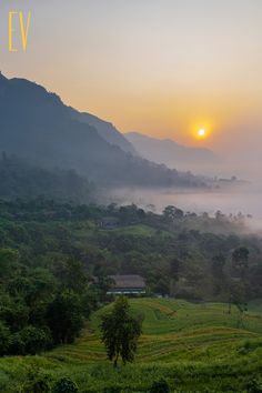 the sun is setting over some hills and trees in the foggy valley with houses on it