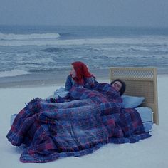 two people laying in the snow under blankets on a bed by the beach with waves coming in