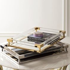 two clear trays with books on them sitting on a marble top table in front of a white wall