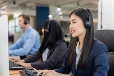 two women wearing headsets are working on their computers in an office setting with other people sitting at desks