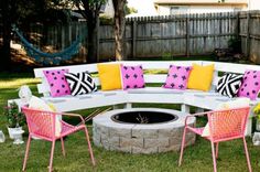 an outdoor fire pit surrounded by pink and yellow pillows, with chairs around the firepit