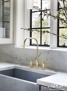 a kitchen sink under a window with marble counter tops and gold faucet handles