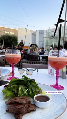 two glasses filled with drinks sitting on top of a table next to steak and salad