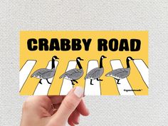 someone holding up a card that says crabby road with three geese crossing the street