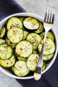 a white bowl filled with cucumbers and topped with seasoning next to a fork