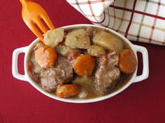 a white bowl filled with meat and potatoes on top of a red table cloth next to a fork