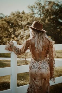 a woman in a dress and hat leaning on a fence with her back to the camera