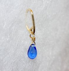 a small blue glass droplet hangs from a gold - plated earring on a white background