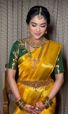 a woman in a yellow and green sari with jewelry on her neck, standing next to a chair