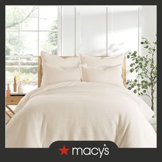 a bed with white sheets and pillows in front of a window, next to a potted plant