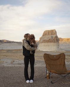 a woman standing next to a camping chair holding a teddy bear in her arms and looking at the desert