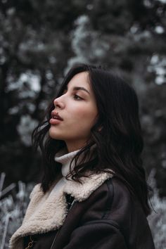 a woman standing in front of some trees wearing a black jacket and she is looking off into the distance