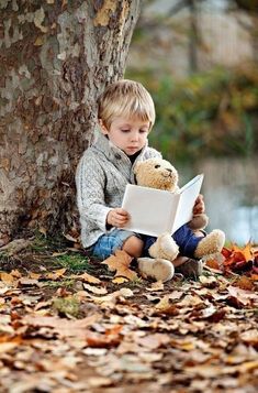 a little boy sitting under a tree reading a book with a teddy bear on his lap