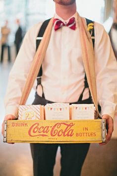 a man in suspenders and bow tie holding a coca - cola crate filled with drinks