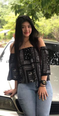 a woman standing in front of a white car wearing jeans and a top with an off the shoulder design