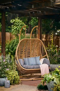 a wicker swing chair with blue cushions in a garden area next to flowers and trees