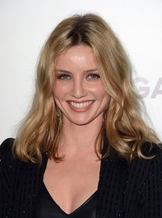 a woman with long blonde hair smiling at the camera and wearing a black top, standing in front of a white wall